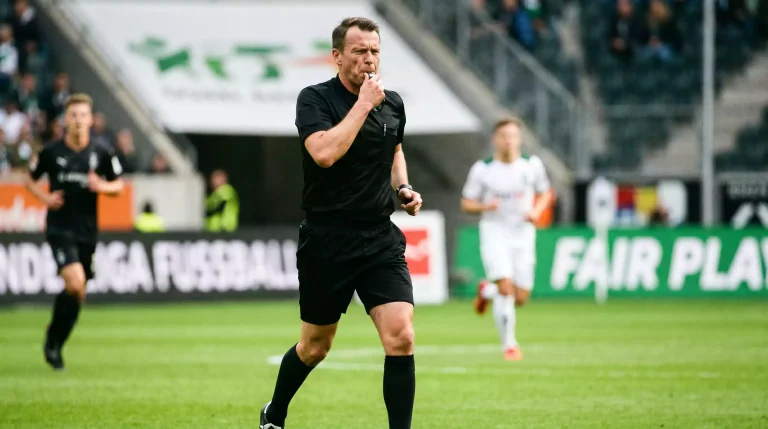 Schiedsrichter in schwarzer Schiedsrichtertracht mit Pfeife auf dem grünen Rasen eines deutschen Bundesliga-Stadions während eines Spiels